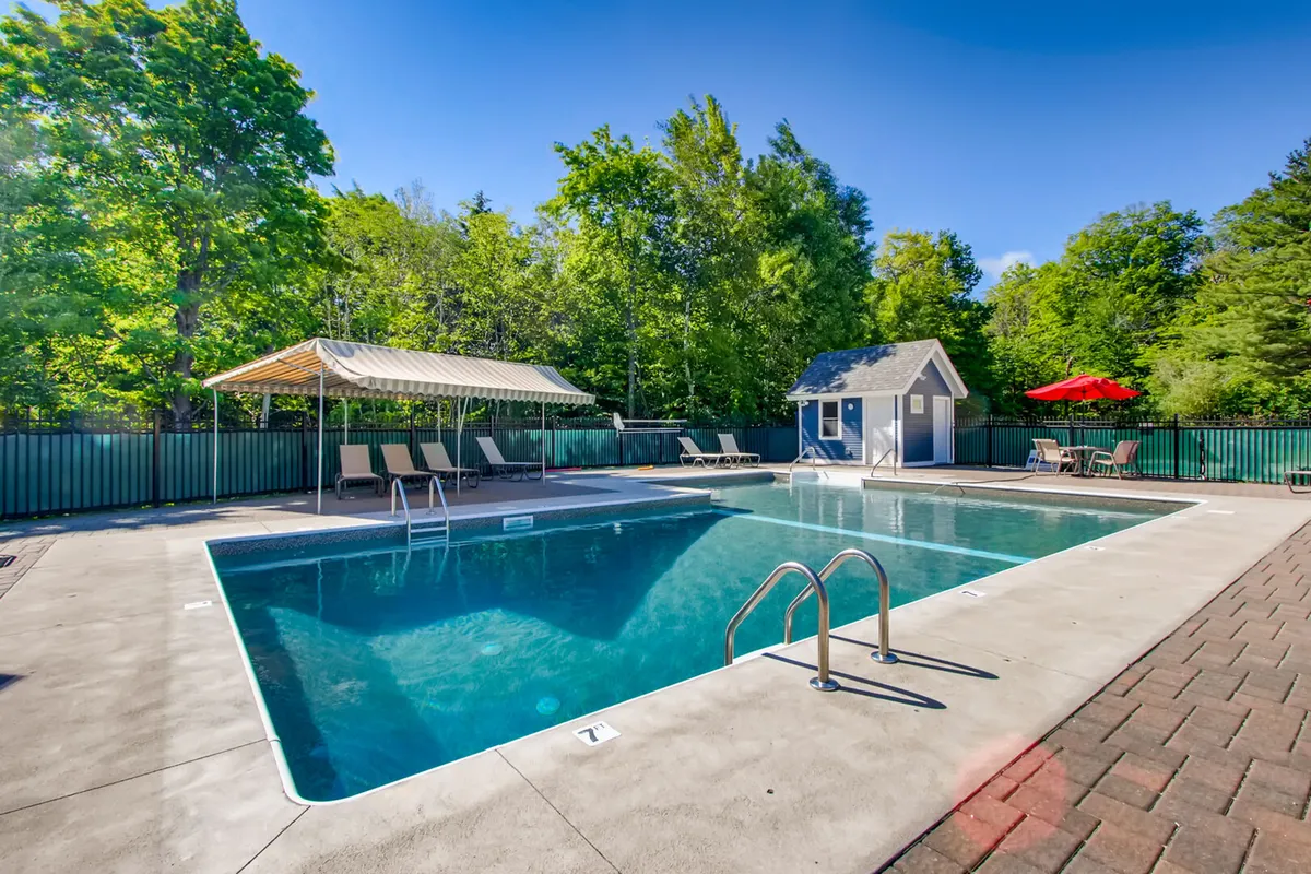 The outdoor swimming pool is surrounded by lounge chairs, a cabana, and a red umbrella, set against a backdrop of greenery and blue sky. The relaxed, inviting pool area is ideal for unwinding after a day spent outdoors.