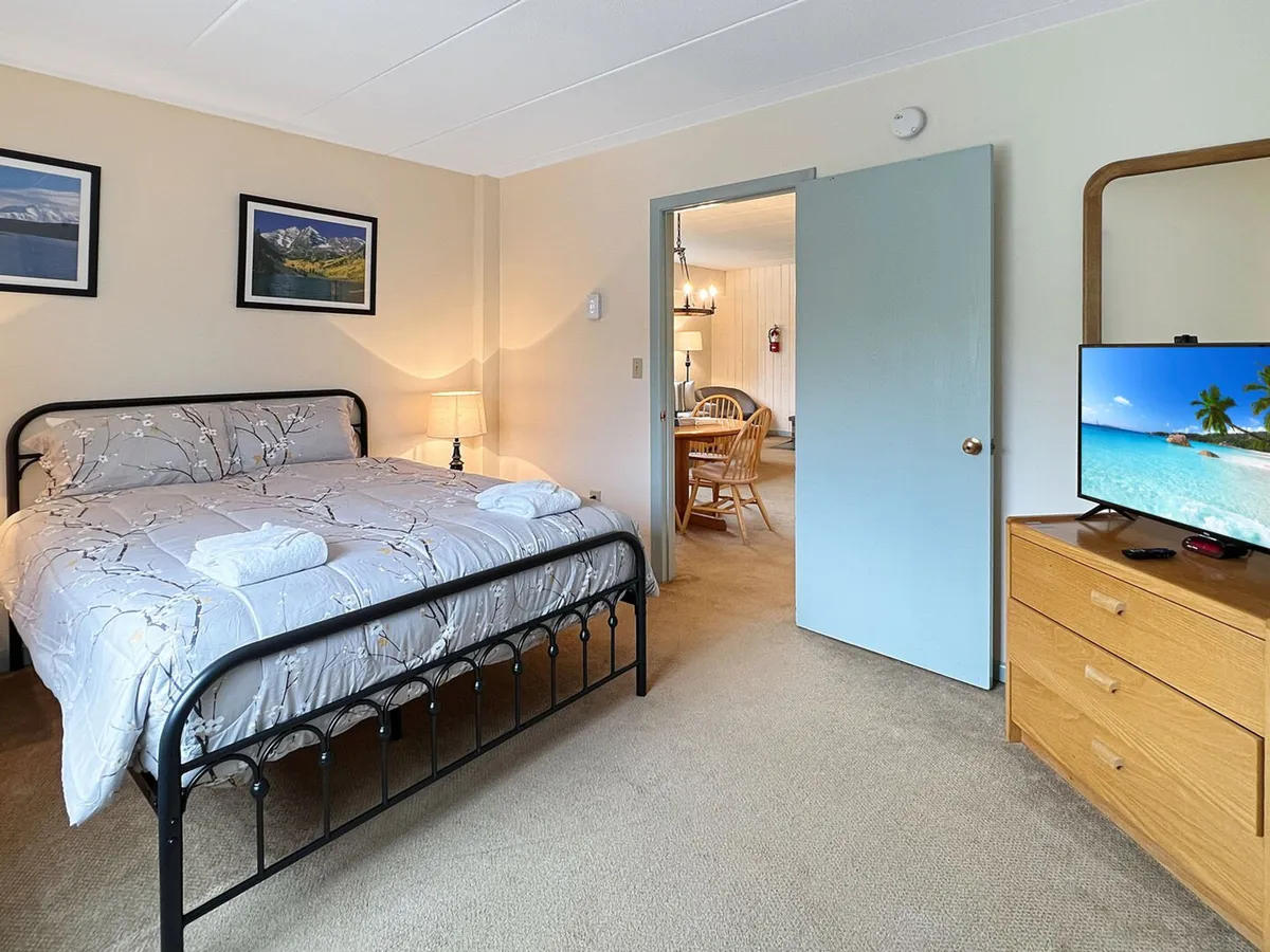 A comfortable bedroom is shown with plush decorative bedding on a queen-size bed. A TV sits on a dresser at the end of the bed, and a blue door leads to an adjacent space, hinting at the proximity of the bathroom. The room feels inviting and restful, perfect for catching up on late-night shows or unwinding after a busy day.