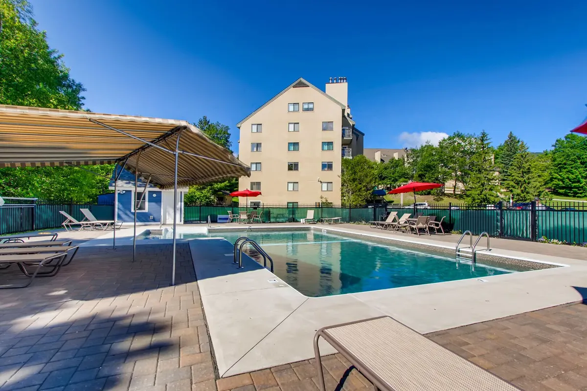 An inviting outdoor swimming pool is surrounded by lounge chairs, vibrant red umbrellas, and green trees, with the resort building facade in the background. The scene is lively and leisure-focused, perfect for family fun under the sun.