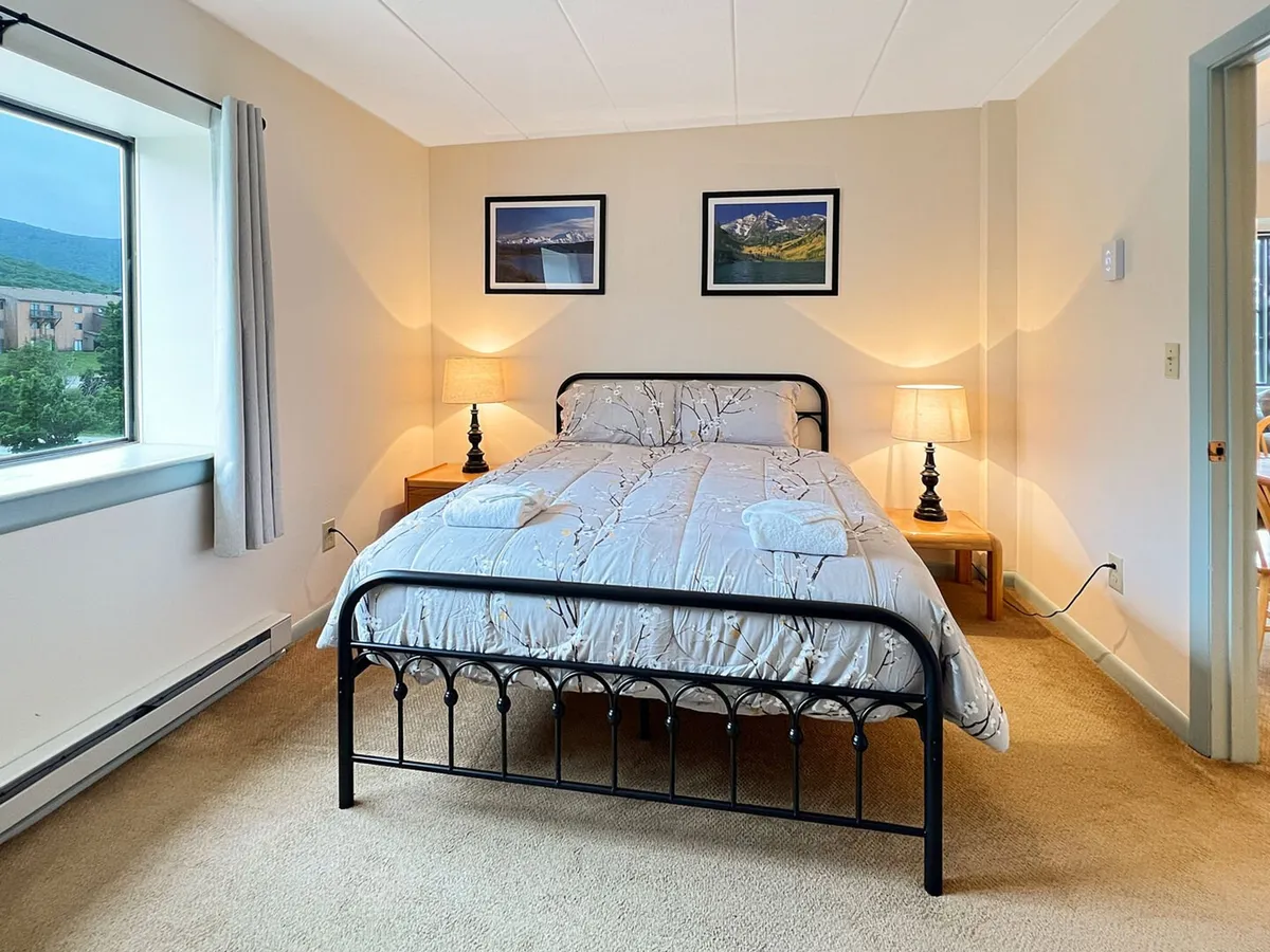 This image features a spacious bedroom with a comfortable queen bed, nightstands, and framed pictures. Large windows offer a beautiful view of the mountains, and the overall atmosphere is warm and inviting, perfect for relaxing after a busy day. The décor has natural, woodsy touches that complement the tranquil vibe of the room.