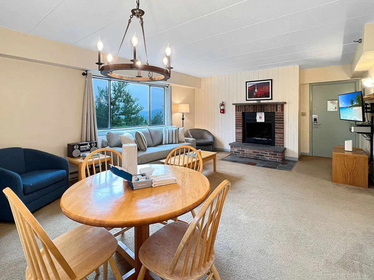 A bright, homey open-concept living/dining area with a comfortable sofa, a large window offering another view of the outdoors, a dining table, and a focal point fireplace. Sunlight fills the room, emphasizing the inviting feel. The fireplace is present but not the center of activity.