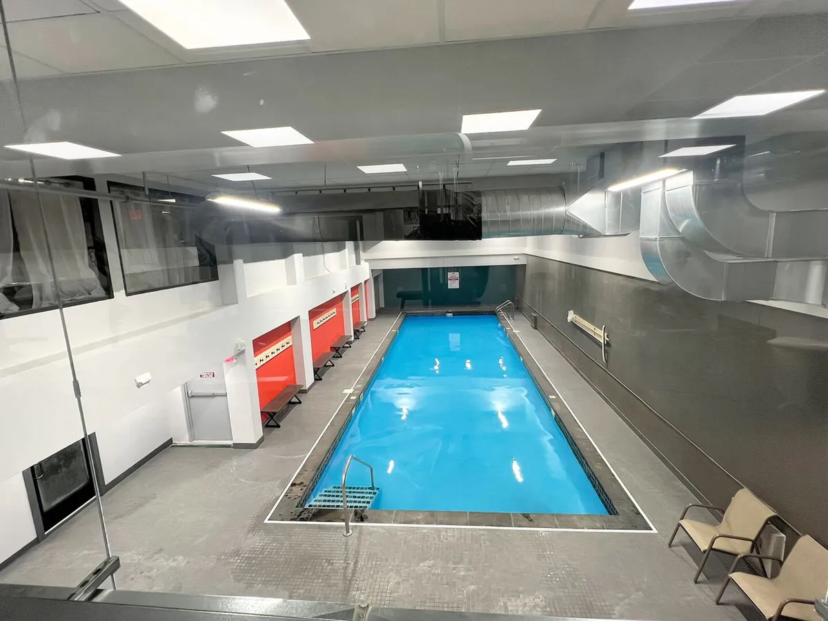 An inviting indoor swimming pool area with glass walls allowing natural light to pour in. Red seating is arranged along one side, and the scene is bright and refreshing. The pool appears large enough for both lap swimming and casual enjoyment.