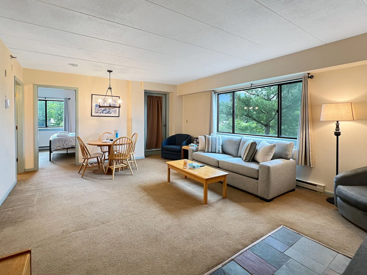 The image features a cozy interior with a comfortable sofa, a dining table set for shared meals, a warm lamp creating inviting lighting, and a window offering views of the surrounding nature. The setup suggests a welcoming space for relaxing, dining, and enjoying time with loved ones after a day on the slopes or golf course.