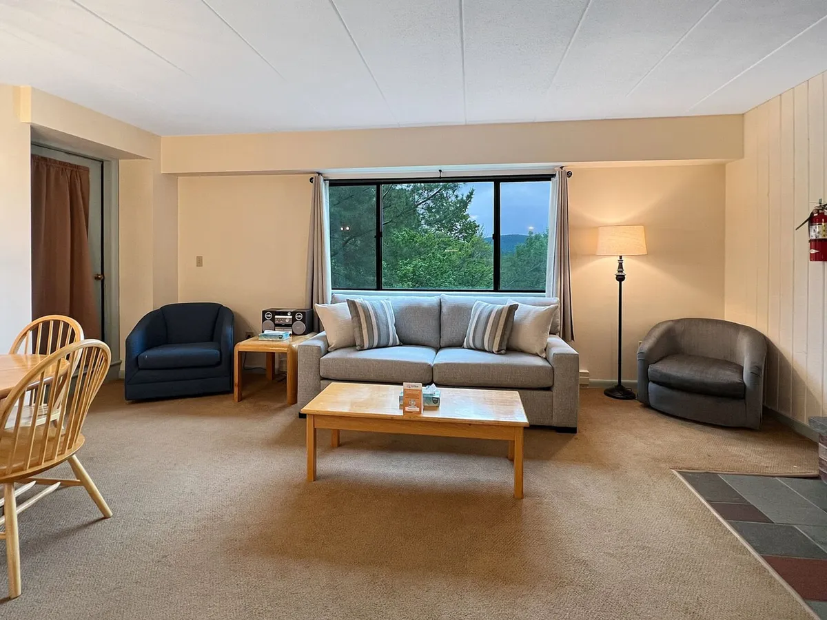 A cozy living room scene featuring a sofa with plush cushions positioned near a large window that frames a sunset view of the surrounding mountains and forests. A dining table is visible, suggesting a space for relaxing, socializing, or game nights. Warm lighting and uncluttered decor create a serene, inviting atmosphere.