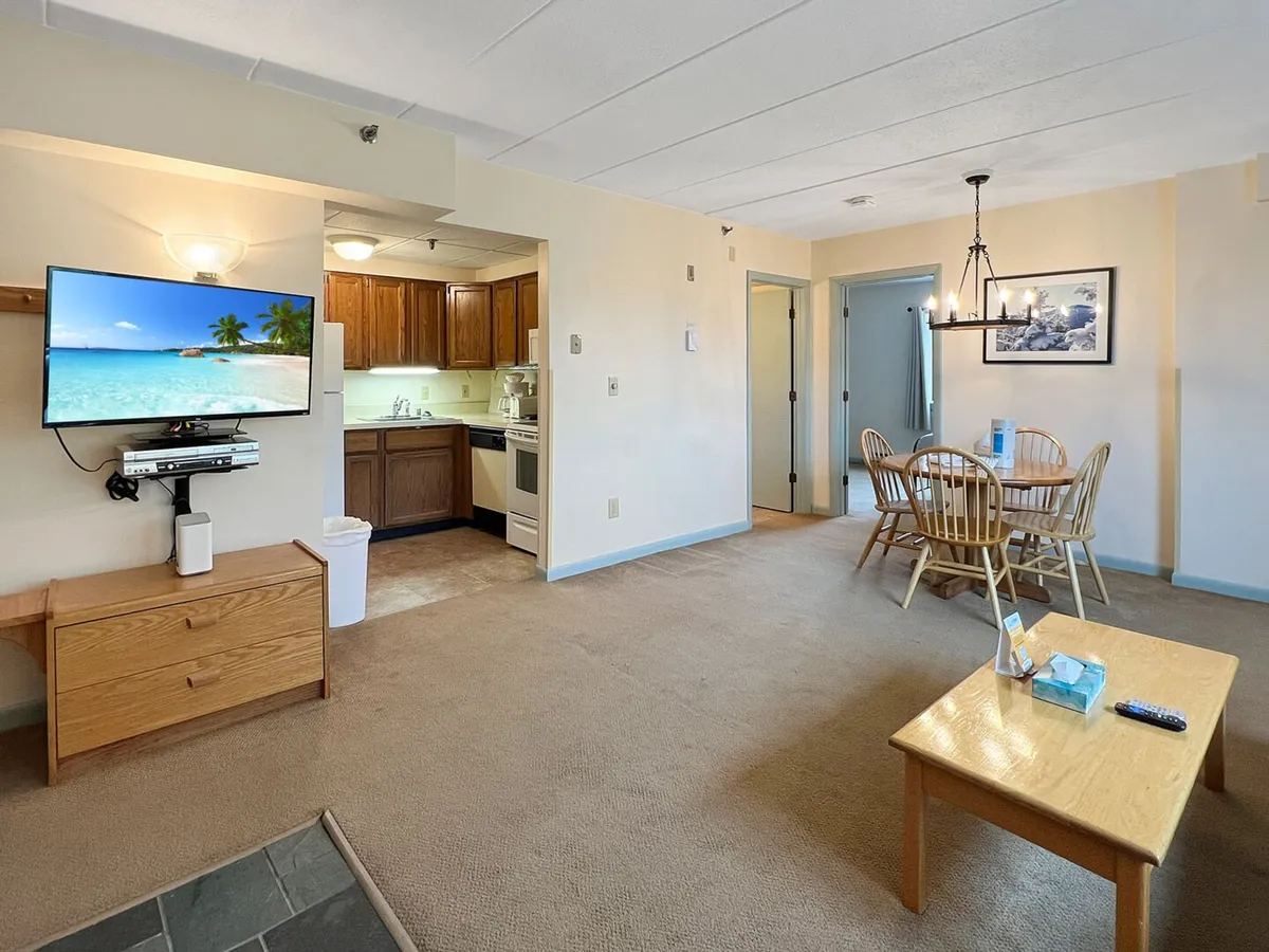 A comfortable interior view featuring a kitchen area, dining table, and a television displaying a beach scene. The space feels relaxed and open, ideal for dining, cooking together, or enjoying entertainment as a group.