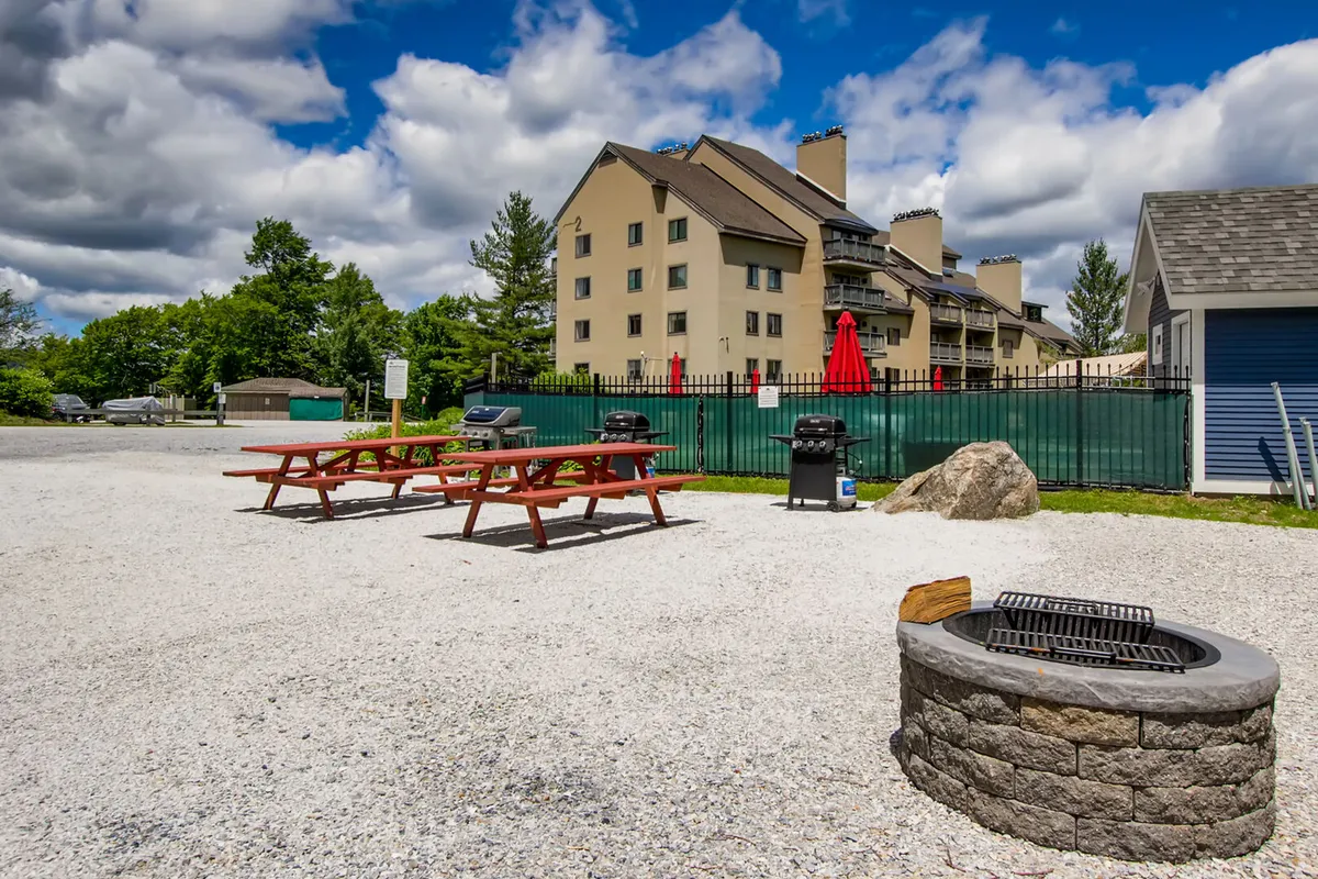 An outdoor gathering space with picnic tables, a fire pit, and grill stations in front of the main building. The area is set up for barbecuing and socializing, reflecting a lively, friendly atmosphere.