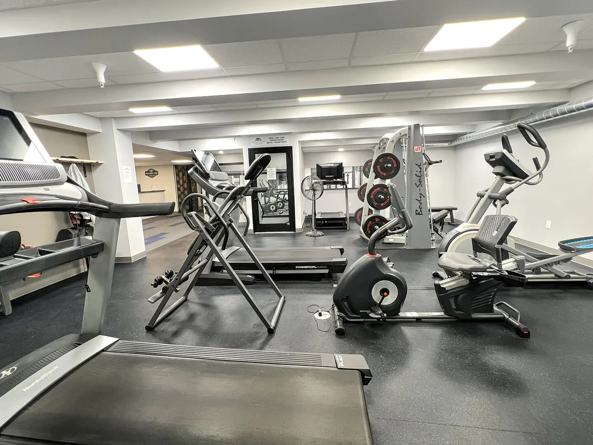A well-equipped fitness center featuring treadmills, an elliptical, exercise bike, weight machine, and a television. The space is clean, well-lit, and geared toward both cardio and strength training.