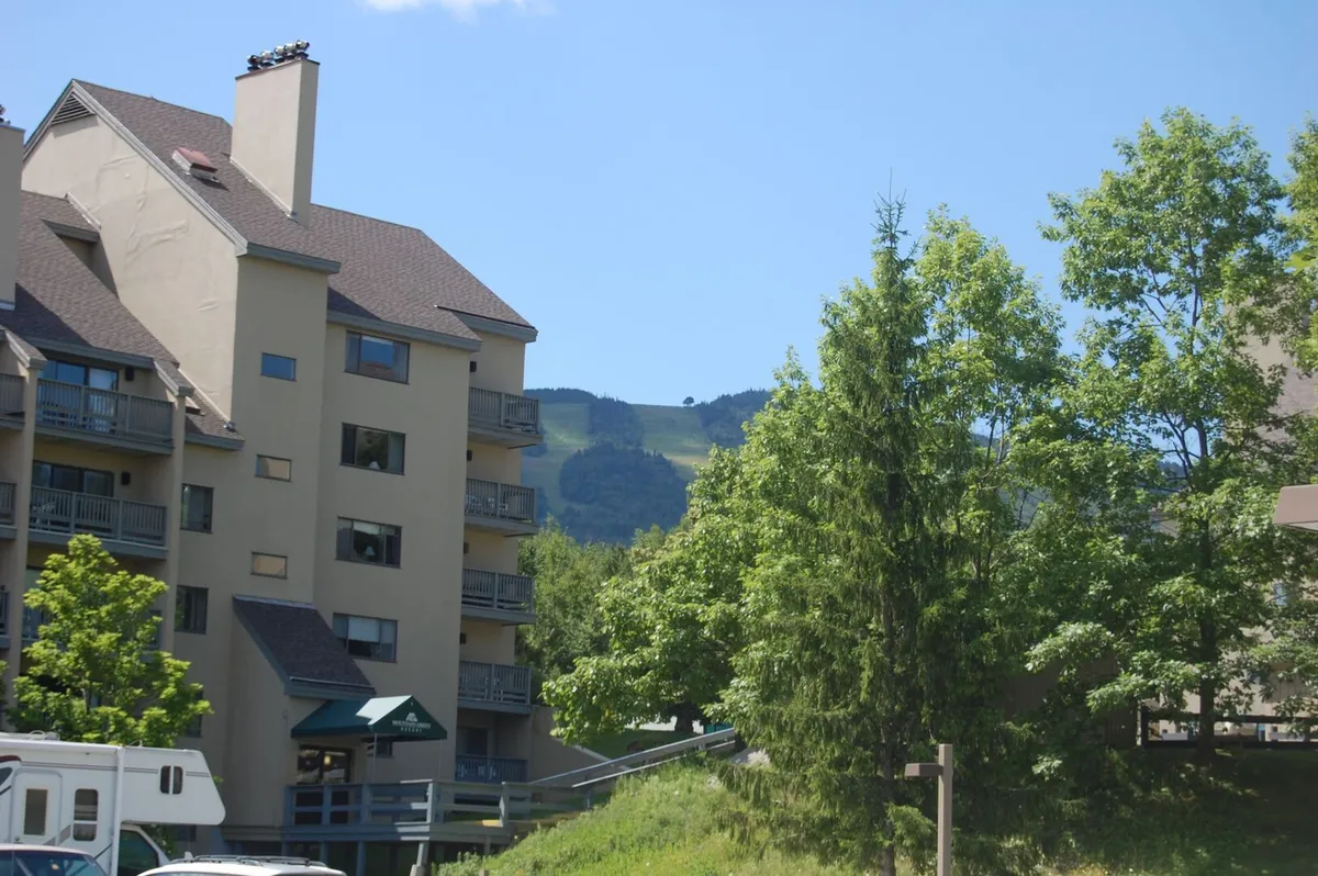 This exterior shot features a multi-story building nestled among green trees with a dramatic mountain backdrop, capturing both the property's proximity to nature and the inviting outdoor environment guests can enjoy in warmer months.