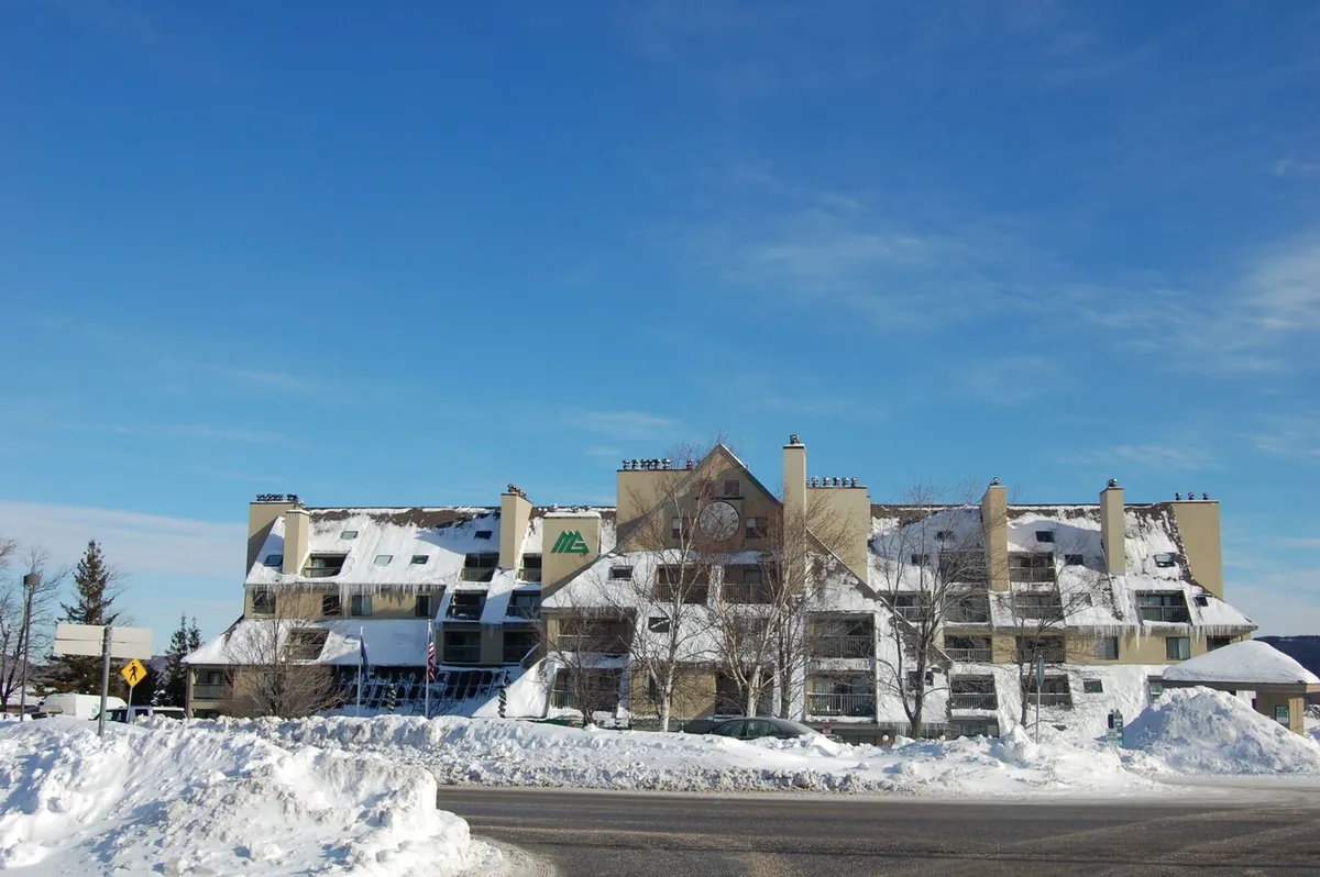A picturesque snow-covered building with icicles hanging from the roof, surrounded by trees blanketed in fresh snow and set against a clear blue sky. The inviting lodge is nestled in a classic Vermont winter landscape, immediately evoking the feeling of a ski resort steps from the slopes.